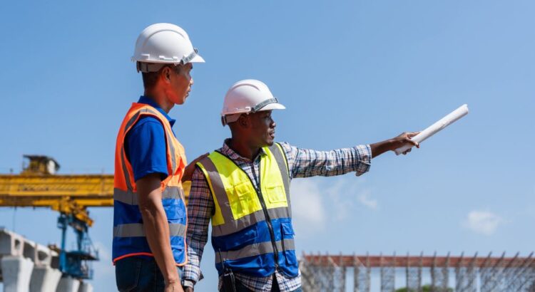 Profissionais da construção civil com capacete e colete de segurança em obra, contexto relacionado às normas e regras da construção civil em atividades técnicas.